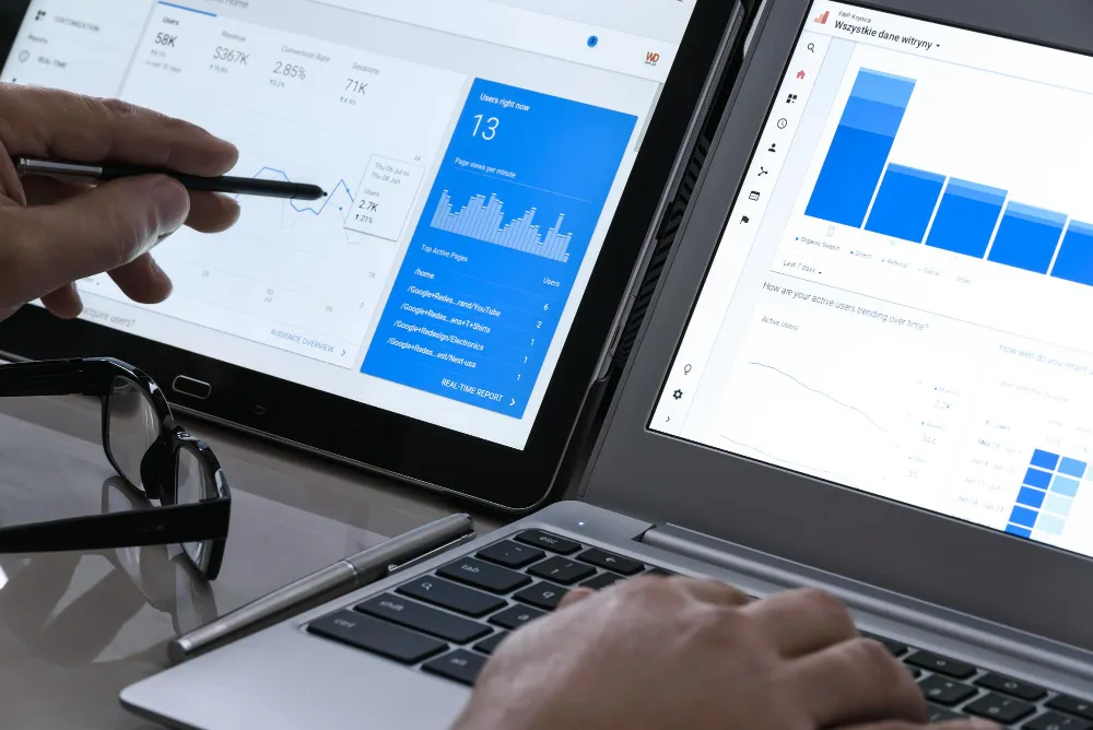 Mãos usando um tablet com uma caneta digital e digitando em um laptop, ambos exibindo dashboards de análise de dados e gráficos, como relatórios em tempo real do Google Analytics. Óculos e uma caneta metálica estão sobre a mesa.