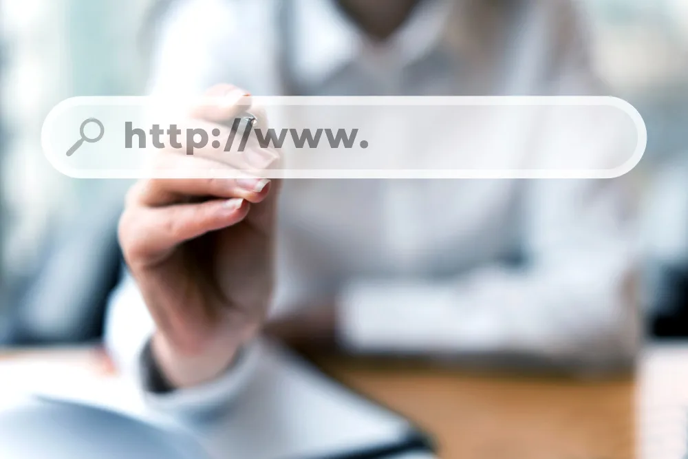 Uma mão apontando para uma barra de pesquisa flutuante com "http://www." preenchido e um ícone de lupa, sobre um fundo borrado que sugere um ambiente de escritório.