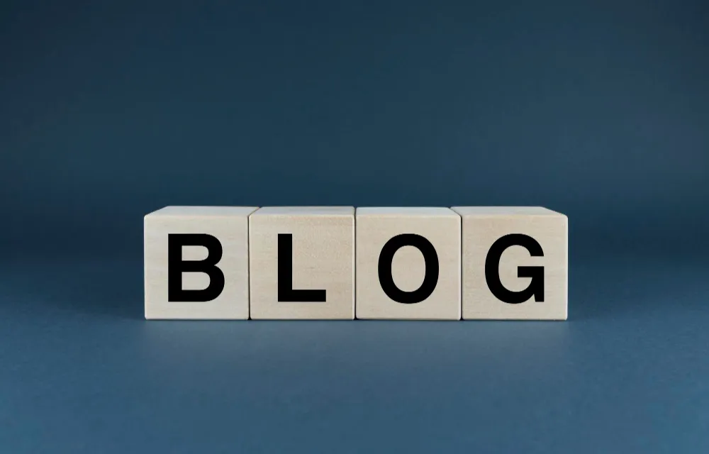 Quatro blocos de madeira alinhados horizontalmente, cada um com uma letra preta formando a palavra "BLOG", sobre um fundo cinza escuro.
