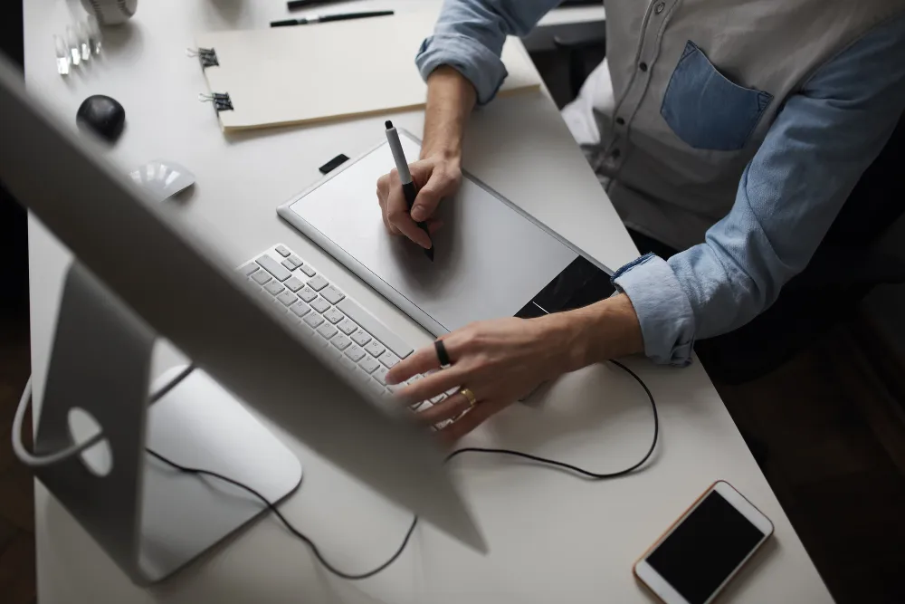 Vista superior de uma pessoa sentada em uma mesa branca, usando uma caneta digital em uma mesa digitalizadora com uma mão e digitando em um teclado com a outra, com um monitor e um celular visíveis.