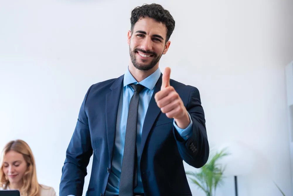 Um homem barbudo e sorridente, vestido com um terno azul e camisa, faz um sinal de "joinha" para a câmera em um escritório, com uma colega de trabalho desfocada ao fundo.