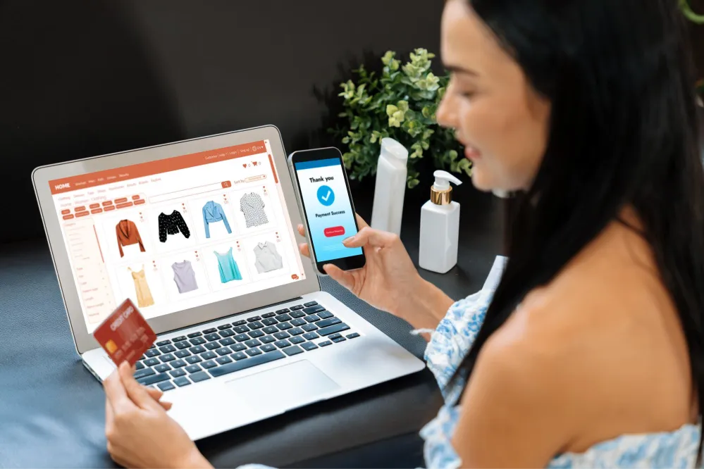 Uma mulher sentada, segurando um cartão de crédito e um smartphone que exibe uma tela de "pagamento bem-sucedido", enquanto olha para um laptop que mostra um site de compras de roupas online.
