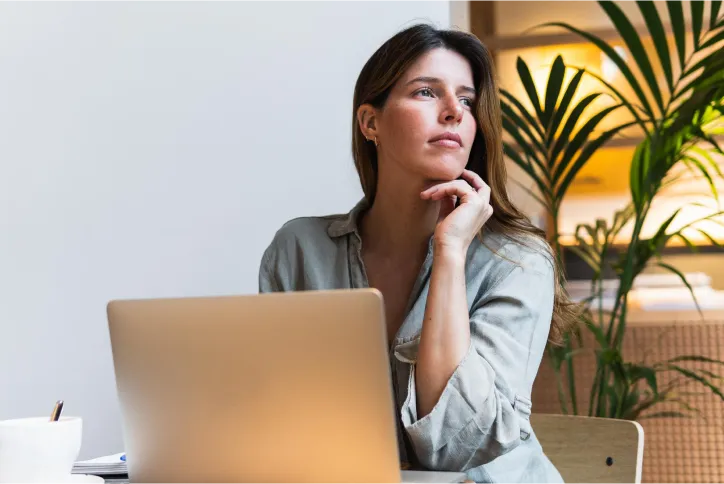 Uma mulher com camisa clara e cabelo castanho comprido, sentada em uma mesa com um laptop à sua frente, com a mão no queixo e olhar pensativo, voltado para o lado direito da imagem. Uma planta verde e uma parede branca estão ao fundo.