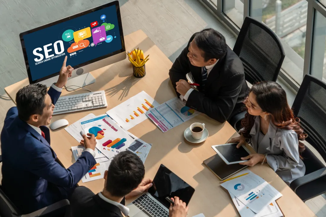 Vista aérea de uma reunião de negócios onde quatro profissionais discutem SEO (Search Engine Optimization), com um monitor exibindo "SEO" e ícones de marketing digital, e gráficos impressos sobre a mesa.