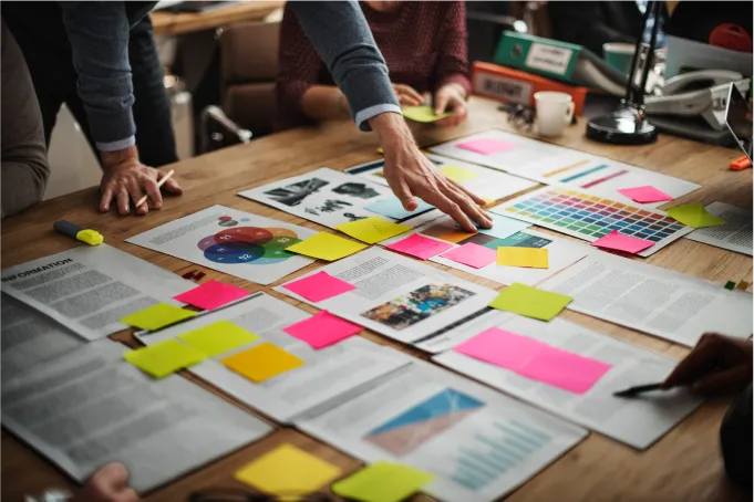 Mesa de trabalho com equipe analisando gráficos, paleta de cores e anotações em post-its durante planejamento de marketing