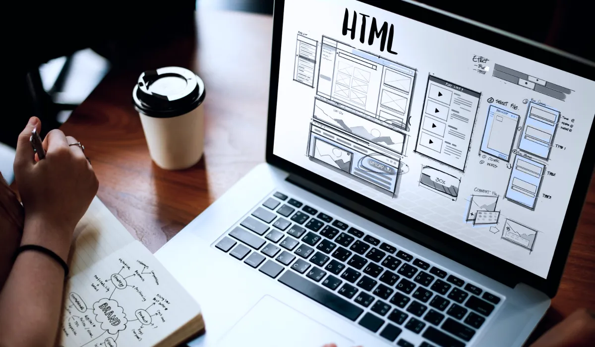 Pessoa planejando estrutura de site em caderno enquanto visualiza wireframe HTML na tela do notebook.