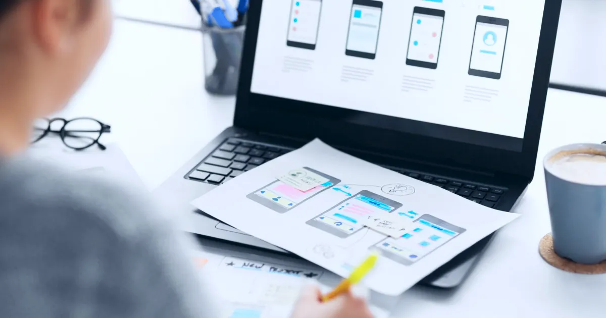 Pessoa desenvolvendo protótipos de aplicativo mobile com rascunhos em papel e tela de laptop.